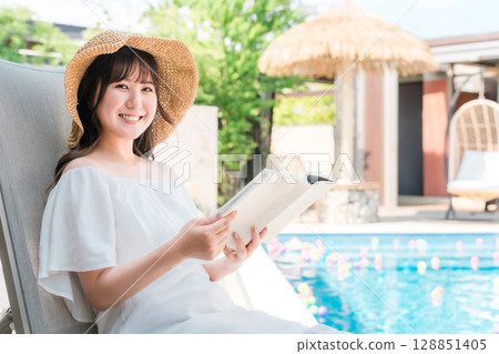 一位年輕的亞洲女性坐在泳池邊的躺椅上看書 一位年輕的亞洲女性坐在泳池邊的躺椅上看書 128851405