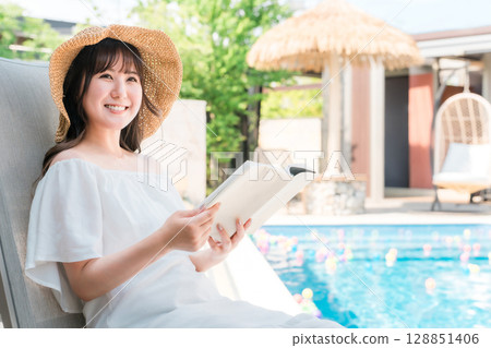 一位年輕的亞洲女性坐在泳池邊的躺椅上看書 一位年輕的亞洲女性坐在泳池邊的躺椅上看書 128851406
