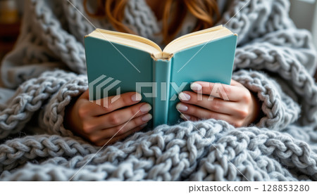 Person wrapped in chunky knit blanket is holding and reading small teal book, creating cozy and calm atmosphere Person wrapped in chunky knit blanket is holding and reading small teal book, creating cozy and calm atmosphere 128853280