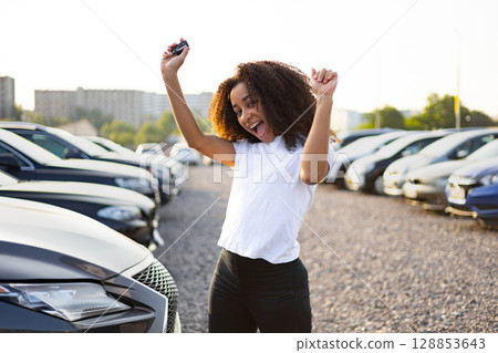 An excited woman celebrates her new car purchase, holding the key high near a car lot with various vehicles. 128853643