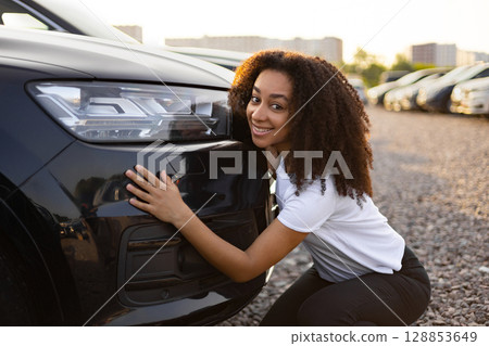 A smiling woman embraces a new black car, likely at a car dealership or lot, excited about her purchase. A smiling woman embraces a new black car, likely at a car dealership or lot, excited about her purchase. 128853649