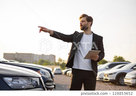 A businessman with a tablet gestures towards a car lot, surrounded by various vehicles on display under a clear sky. A businessman with a tablet gestures towards a car lot, surrounded by various vehicles on display under a clear sky. 128853652