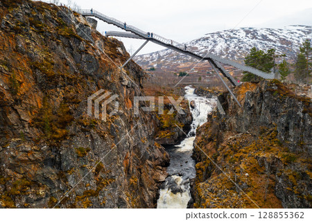 Impressive Voringsfossen footbridge spans over deep gorge with winding river against snowy mountains. Dramatic natural landscape in Norway region Impressive Voringsfossen footbridge spans over deep gorge with winding river against snowy mountains. Dramatic natural landscape in Norway region 128855362