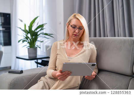 Relaxed mature blonde woman in casual outfit browsing on digital tablet while sitting comfortably on couch in bright living room Relaxed mature blonde woman in casual outfit browsing on digital tablet while sitting comfortably on couch in bright living room 128855363