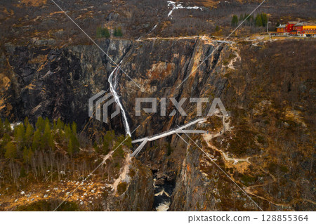 Voringsfossen waterfall cascades down rugged dark rock face with modern bridge crossing over canyon. Dramatic scale of natural wonder in Norway 128855364