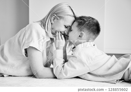 Little son showing love to his mom. Young mother and her kid lying on white carpet and spending time together at home. Lovely afternoon with family. Black and white image. 128855602