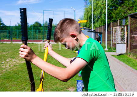 Boy exercising on outdoor fitness equipment in a park on a sunny day. 128856139