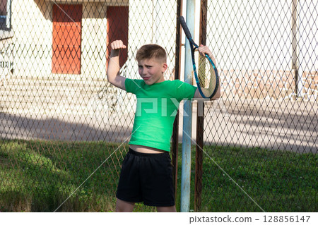 Young boy shows off his muscles after a tennis match. He leans against the fence with his racquet. 128856147