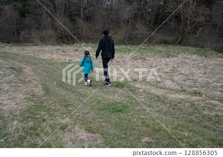 Child and adult embrace nature on a tranquil pathway surrounded by lush greenery in early spring, capturing the joy of exploration and togetherness in a picturesque landscape 128856305