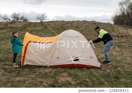 Setting up a cozy tent on a grassy hillside under a cloudy sky, with a child eagerly helping while enjoying outdoor adventure together Setting up a cozy tent on a grassy hillside under a cloudy sky, with a child eagerly helping while enjoying outdoor adventure together 128856411