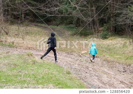 Two figures traverse a muddy forest trail surrounded by trees in a tranquil outdoor setting during a cool day, evoking a sense of adventure and exploration 128856420