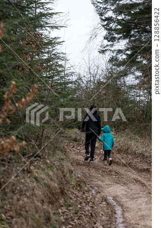 Walking hand in hand along a forest trail during a chilly afternoon, a parent and child explore nature, surrounded by tall trees and earthy tones Walking hand in hand along a forest trail during a chilly afternoon, a parent and child explore nature, surrounded by tall trees and earthy tones 128856422