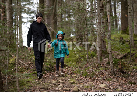Walking through a tranquil forest path during early spring, a young boy holds hands with a girl dressed in a bright jacket as they explore nature together 128856424