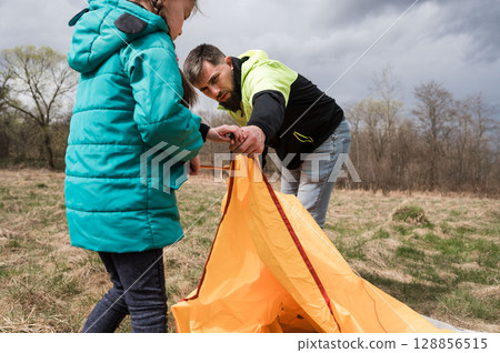 Family camping adventure unfolds as a father and daughter set up an orange tent under a moody sky in the great outdoors during springtime exploration Family camping adventure unfolds as a father and daughter set up an orange tent under a moody sky in the great outdoors during springtime exploration 128856515