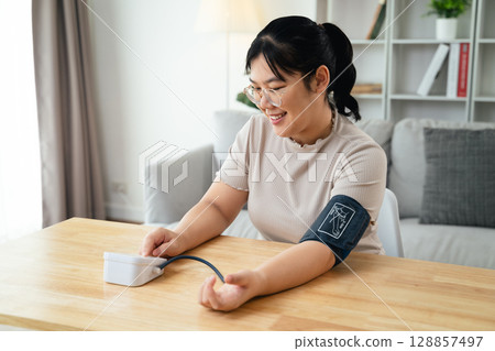 An Asian woman uses a portable blood pressure monitor in her living room. Health awareness concept. An Asian woman uses a portable blood pressure monitor in her living room. Health awareness concept. 128857497