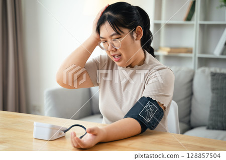 An Asian woman uses a portable blood pressure monitor in her living room. Health awareness concept. 128857504
