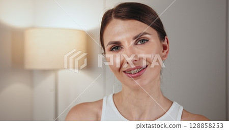 Portrait of a young Caucasian woman wearing braces, smiling warmly in home interior. Represents dental care and orthodontics. Bright, inviting atmosphere with soft lighting in background. 128858253