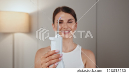 Young Caucasian woman with braces using dental irrigator showcasing oral health care tool at home. Young Caucasian woman with braces using dental irrigator showcasing oral health care tool at home. 128858280