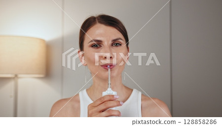 Caucasian young woman with braces using dental irrigator in home setting. Promoting good oral hygiene and dental care routine. She appears focused and attentive. 128858286