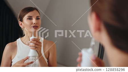 Young Caucasian woman with brown hair using water irrigator for dental care. Standing in front of mirror, maintaining oral hygiene with braces. Casual indoor bathroom setting. Young Caucasian woman with brown hair using water irrigator for dental care. Standing in front of mirror, maintaining oral hygiene with braces. Casual indoor bathroom setting. 128858296