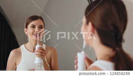 Young Caucasian woman with braces using dental irrigator in bathroom, smiling and looking in mirror for oral hygiene care. 128858299