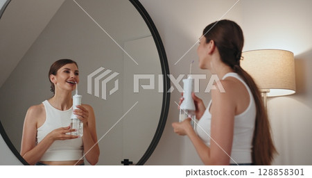 Young Caucasian woman with braces using water irrigator for dental hygiene in front of mirror, smiling and wearing casual tank top, relaxed atmosphere. 128858301
