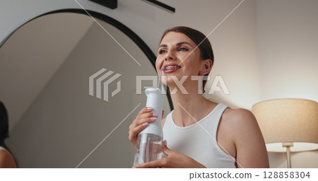 Young Caucasian woman smiling while using dental irrigator for oral hygiene. Braces visible as she looks in mirror. Promotes good dental care routine. 128858304