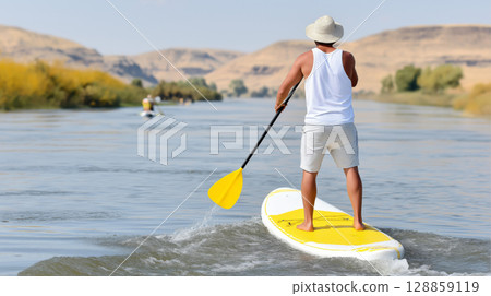 Young male paddleboarding on calm river surrounded by scenic hills, rear view 128859119