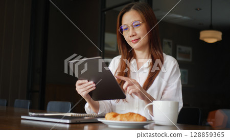 side view of Asian woman wearing eyeglasses using tablet for work in the morning on the wooden dining table, working from home concept side view of Asian woman wearing eyeglasses using tablet for work in the morning on the wooden dining table, working from home concept 128859365
