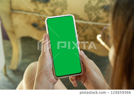 Asian woman with eyeglasses holding smartphone in hand, phone mockup with blank empty green screen template using cell phone relaxing on sofa at home. over shoulder view. mobile apps advertising Asian woman with eyeglasses holding smartphone in hand, phone mockup with blank empty green screen template using cell phone relaxing on sofa at home. over shoulder view. mobile apps advertising 128859366