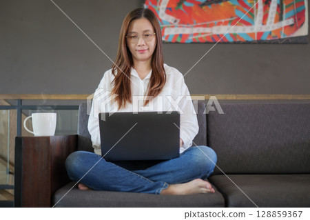 front view of Asian woman with eyeglasses sitting on sofa, using laptop computer for browsing and searching online on the internet, working from home in the morning with hot coffee. remote location front view of Asian woman with eyeglasses sitting on sofa, using laptop computer for browsing and searching online on the internet, working from home in the morning with hot coffee. remote location 128859367