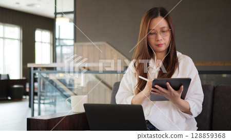 Asian woman using laptop for remote working at home or coffee shop, using earphones for conference and writing note on digital tablet. learning training, studying online seminar or video calling. Asian woman using laptop for remote working at home or coffee shop, using earphones for conference and writing note on digital tablet. learning training, studying online seminar or video calling. 128859368