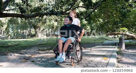 Young Woman Assisting Man in Wheelchair Enjoying a Stroll in a Sunny Park Surrounded by Lush Greenery and Trees Young Woman Assisting Man in Wheelchair Enjoying a Stroll in a Sunny Park Surrounded by Lush Greenery and Trees 128859968