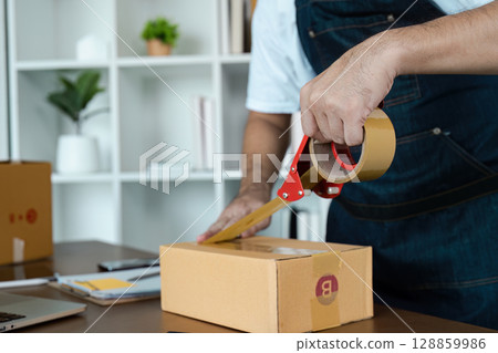 Man Sealing Cardboard Box with Tape in Home Office Setting for E-commerce or Small Business Packaging and Shipping 128859986