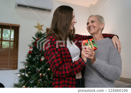 Daughter hug elderly mother with gift box at home in front of tree decorated with Christmas decorations Daughter hug elderly mother with gift box at home in front of tree decorated with Christmas decorations 128860106