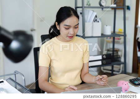 Young Woman Engaged in Financial Planning and Savings Management at Home Office with Laptop, Credit Card, and Piggy Bank 128860295