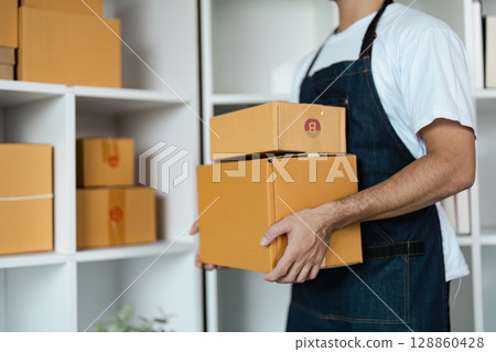 Entrepreneur in Apron Holding Cardboard Boxes in Small Business Warehouse Setting 128860428