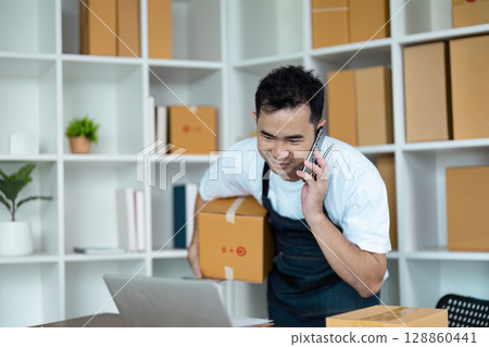Young Entrepreneur Managing Online Orders in a Modern Small Business Office with Packaging Boxes and Laptop Young Entrepreneur Managing Online Orders in a Modern Small Business Office with Packaging Boxes and Laptop 128860441