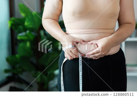 Person Measuring Waist with Tape in Modern Home Setting Highlighting Overweight and Health Awareness Person Measuring Waist with Tape in Modern Home Setting Highlighting Overweight and Health Awareness 128860511