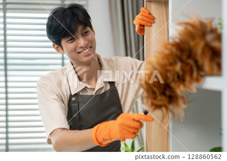 Smiling Housekeeper Dusting Shelves in Modern Home with Feather Duster and Protective Gloves Smiling Housekeeper Dusting Shelves in Modern Home with Feather Duster and Protective Gloves 128860562