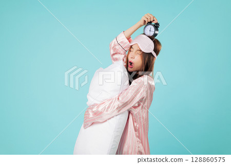 Tired woman in pajama with sleep mask embraces pillow, holding alarm clock above her head while yawning, as she hates when the clock rings and wakes her up, headshots isolated studio blue background Tired woman in pajama with sleep mask embraces pillow, holding alarm clock above her head while yawning, as she hates when the clock rings and wakes her up, headshots isolated studio blue background 128860575