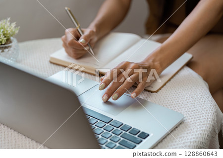 Woman Writing in Notebook While Using Laptop at Home Office for Remote Work or Study in Cozy Environment 128860643
