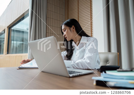 Professional Businesswoman analysis Financial Reports in a Modern Office Setting with Laptop and Document Professional Businesswoman analysis Financial Reports in a Modern Office Setting with Laptop and Document 128860706