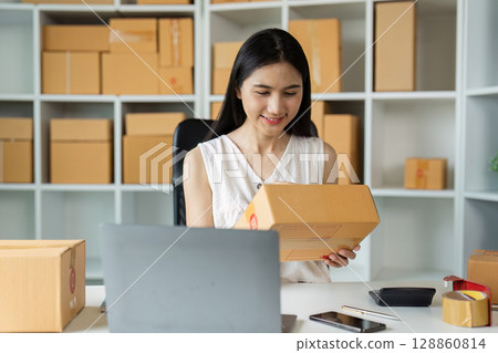 Young Entrepreneur Working on Small Business Packaging Orders in Home Office with Laptop and Smartphone 128860814