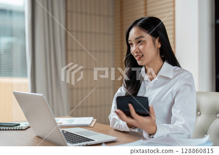 Professional Woman Working from Home Office with Laptop and Smartphone, Smiling and Engaged in Business Tasks Professional Woman Working from Home Office with Laptop and Smartphone, Smiling and Engaged in Business Tasks 128860815