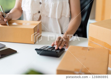 Small Business Owner Calculating finance and Managing Online Order with Cardboard Boxes on Desk 128860891