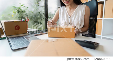 Woman Preparing Packages for Online Small Business with Laptop and Calculator on Desk Woman Preparing Packages for Online Small Business with Laptop and Calculator on Desk 128860982