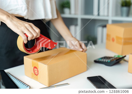 Entrepreneur Packing Orders for Online Business with Cardboard Boxes, Tape, and Calculator in Modern Office Entrepreneur Packing Orders for Online Business with Cardboard Boxes, Tape, and Calculator in Modern Office 128861006