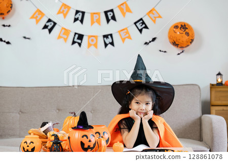 Cute Child in Witch Costume Celebrating Halloween with Decorations and Pumpkins Indoors 128861078
