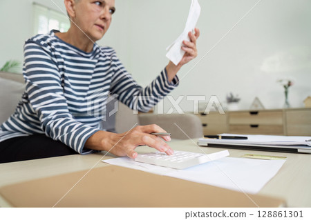 Senior Woman Managing Finances at Home with Calculator and Documents in Living Room Senior Woman Managing Finances at Home with Calculator and Documents in Living Room 128861301
